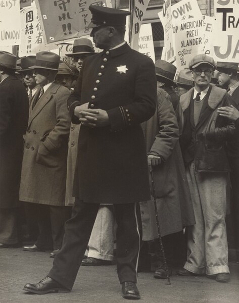 A uniformed policeman standing in front of a dense crowd of men holding protest signs in English and Japanese nearly fills the height of this vertical black and white photograph. The policeman either has a ruddy complexion or his face might be in shadow. He stands with his body facing us but he turns his head to our left in profile. His hands are crossed over his chest and one thumb is hooked into his jacket between two of the eight brass buttons down the front. A seven-pointed star reflects light on his chest and his flat-topped hat has a short, shiny brim angled down over his eyes. The jacket comes to his knees and his pants are lined with gold or other light material down the side. His feet are widely planted, and he wears dark shoes. The men in the crowd behind appear to be light skinned. Many wear button-down, collared shirts with ties tucked into long coats. Many also wear fedora hats and look off to our left, the same direction as the policeman. One man to our right wearing a floppy cap and glasses looks out at us with his hands in his jacket pockets. Some of the legible English words on the protest signs read “UNION,” “AMERICAN PRESS SLANDER against the,” and “JA.”