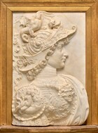 Set within a gold frame, the head and chest of a young man wearing an ornate helmet and armor, facing our right in profile, is carved in relief from white marble in this vertical panel. The man has a long nose, smooth cheeks, closed bow lips, and a snub chin. Curls burst from beneath his helmet, which sweeps from up over his forehead down to the back of his neck. The lifted visor curves up and out past his nose, and is decorated with vines and flowers. The crown of the helmet spirals like a snail shell over the man’s head, and a dragon perches along the top. The dragon has a dog’s head, a scaled body, wings like a bat, a serpent-like tail, and a section of its long neck has been broken off. Two crimped ribbons flutter up and behind the man’s head from the back of the helmet, where it covers his neck. His chest is covered to his neck with a highly ornamented breast plate. At the front of the chest, an adult face with brows furrowed and mouth wide open is flanked by wings. The shoulder we can see is covered with a medallion showing a nude woman holding a cornucopia and a merman. Beneath the medallion, a row of six faces, each on its own narrow panel, fan out over the man’s upper arm. Scrolling vines, leaves, and flowers fill the remaining space around the medallion and winged head. A few pale, parchment-white veins run through the surface of the marble. The panel sits on a shallow, trapezoidal ledge and is framed in a stepped gold frame on the remaining three sides.