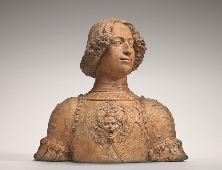 This freestanding, beige, terracotta bust shows the head and shoulders of a cleanshaven man, Giuliano de' Medici, wearing a suit of armor. The bust is straight across the bottom so it rests directly on the white surface on which it is photographed. In this view, the man’s shoulders are square to us, and he looks off to our right, chin lifted. His eyes are blank, and he has a bumped, aquiline nose and rounded cheeks. His lips are closed, the corners slightly pulled back. His thick, curly hair is cut to create an angle down from his eyebrows, on either side of his forehead, to the back of his neck. His breastplate has a high collar, and, at the front center, a man’s head is sculpted in relief with his mouth wide open, head flanked by upswept wings. The yelling man’s teeth and tongue are visible, and he has wavy hair and prominent ears. The pauldrons, the armor covering Giuliano's shoulders, and the rest of the breastplate are decorated geometric shapes and curved lines that end in sharp points.