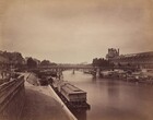 We seem to stand on a paved walkway running alongside a river as we look across the placid water towards a bridge spanning two banks of an elegant city in this horizontal sepia-toned photograph. Printed in warm, rich tones of brown and ivory, the horizon line comes almost halfway up the composition. A few low, blocky barges and boats line the river in front of us and along the opposite bank. The span of the bridge ahead of us is supported by three shallow arches with descending circles where the arches meet the pillars and flat top of the bridge deck. A long building with rows of windows leading to a high peaked roof runs parallel to the river to our right. More buildings face the river to our left in the distance. A few shadowy forms near the left edge of the photograph, close to us, were created as people moved during the long exposure time needed for the image.