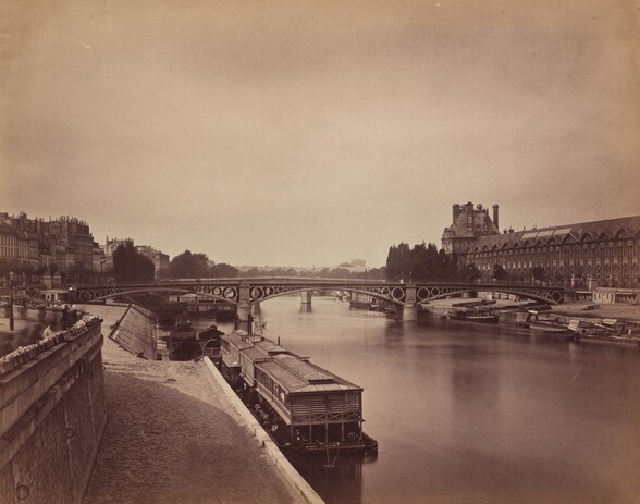 From just over a paved walkway running alongside a river, we look across the placid water toward a bridge spanning two banks of an elegant city in this horizontal, sepia-toned photograph. Printed in warm, rich tones of brown and ivory, the horizon line comes almost halfway up the composition. A few low, blocky barges and boats line the smooth river in front of us and along the opposite bank. The span of the bridge ahead of us is supported by three shallow arches with circles in descending sizes where the arches meet the pillars and the flat top of the bridge deck. A long building with rows of windows leading to a high peaked roof runs parallel to the river to our right. More buildings face the river to our left in the distance. A few shadowy forms near the left edge of the photograph, close to us, were created as people moved during the long exposure time needed for the image.