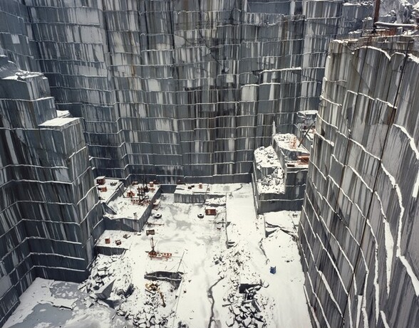 We look down into a straight-sided quarry at mining equipment that appears miniscule in scale in this nearly square color photograph. The walls are streaked with pale and dark gray, and snow rests on narrow ledges between the quarried levels and on the ground. At first glance we might think we’re looking down the sides of skyscrapers onto a city block.