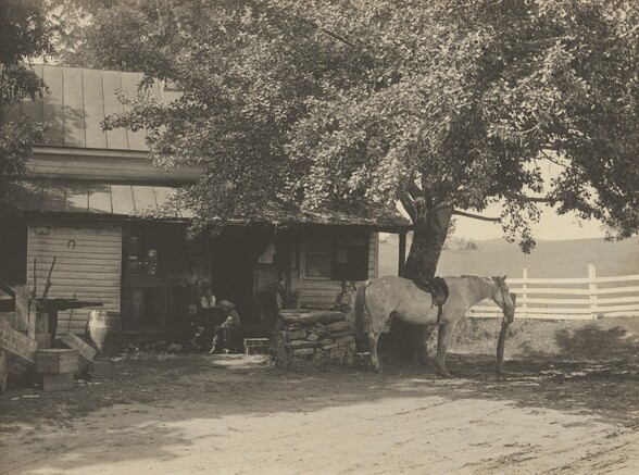Six people sit or stand in the shade of a porch on a building off a dirt road in this horizontal black and white photograph. A saddled white horse stands next to a hitching post near a tree with a sprawling canopy to our right. A man smoking a pipe on the porch sits under a paper bulletin curling away from where it was posted on the wall behind him. Three children sit near some steps perhaps playing a game. An upside-down horseshoe hangs on the horizontal siding to our left. Wood boxes, a barrel, and other equipment are piled nearby.
