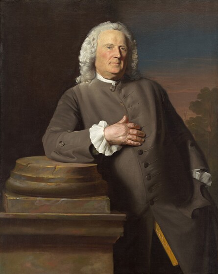 Shown from the knees up, and older, clean-shaven, pale-skinned man rests one elbow on a broken column in this vertical portrait painting. He rests on his right elbow, on our left, so his body creates a diagonal from the lower right to the top center of the composition. He looks at us with clear blue eyes under low brows. His fleshy cheeks are lightly flushed. Wrinkles line his mouth, which is closed in a slight smile. He has jowls and a double chin, and his face is marked with few flesh-colored bumps near his mouth and under one eye. Thick, white hair is brushed back from his forehead, and shoulder-length curls frame his wide face. His is lit from our left so the far side of his face is largely in shadow except for that eye. A strip of bright white fabric encircles his neck above the collar of his long, steel-gray coat. Only the bottom half of the buttons down the front are fastened. White ruffles line his wrists. A glimpse of a golden yellow garment and dark trousers are visible where the coat splits near the bottom of the painting. His left hand, to our right, is thrust into that coat pocket. The other elbow rests on the light brown stone column base on a tall pedestal, and he touches his midsection with those fingers. The left half of the background is dark brown and the right half opens onto a green tree silhouetted against a hazy-pink and aquamarine-blue sky.