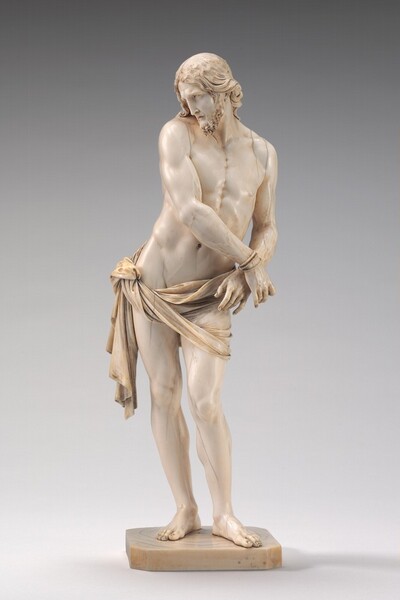 This carved ivory statue shows a mostly nude man standing on a square base with his hands bound to one side as he looks over the other shoulder. In this photograph, his wrists are tied together to our right, and he looks off to our left with his face tucked close to that shoulder. He has a curly beard and long wavy hair, which is pushed back over his shoulders. His lips are parted and his brows gathered. Both feet are planted on the base but most of his weight rests on his straight right leg, on our left, and the other knee is bent. This makes his right hip, on our left, jut out as his torso angles to our right. A loincloth drapes around his hips and ties at his right hip, on our left. His torso, arms, and legs are muscled, and veins stand out on his hands and arms. Some cracks are visible along the surface.