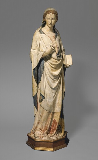 This wooden, painted, free-standing sculpture is of a young woman standing and holding an open book with one hand and holding the other to her chest. The woman’s skin, gown, and long mantle are painted cream white. The mantle covers the back of her head and has a denim-blue lining visible in the folds around her shoulders, waist, and inside the hanging sleeves. It falls just below her knees and wraps around her long gown. The skirt below her knees has traces of brick red but much of the pigment there is lost. In this photograh, her body is angled to our right but she turns her head to look down in front of us with pale brown eyes. Wavy, brown hair is parted down the middle to frame her face. Her right hand, on our left, is raised to touch her chest while her other hand holds an open book at her waist, its pages facing outward. She stands on an octagonal wooden base painted gold. The surface of the sculpture is cracked, flaking, and worn in some areas, allowing the underlying wood to show through.