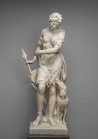 Carved from white marble, a young woman holds a short spear by one side and looks down toward a dog standing by her other side in this free-standing sculpture. Her body faces us in this photograph, and her head is tipped forward and to our right. She has a straight, delicate nose, and her thin lips are parted, possibly in a slight smile. Her wavy hair is held back under a band with an ornament on the crown, and a long curl falls behind one shoulder. Her knee-length garment exposes one firm breast and half of the other. The drapery clings tightly to her body and legs so the folds accentuate her curves. A longer garment falls from her waist, behind her legs, to pool at her sandaled feet. She stands with her weight mostly on her left leg, to our right, and the other knee bent. She holds the shaft of the spear with her right hand, to our left. Her other arm is drawn across her body at waist level. With that hand, she loosely holds the dog’s leash, which then falls across her thighs to the dog, sitting at her feet to our right. The short haired, thin, muscular hound is seated so its chest squares toward us. It twists its head up to look at the woman, its nose pressed along her leg. Its mouth is open, and the tongue curls out over sharp teeth. The dog wears a snug, buckled collar. The woman and dog stand on a square platform of white marble resting on a slab veined with charcoal gray and black, against a smoke-gray wall.