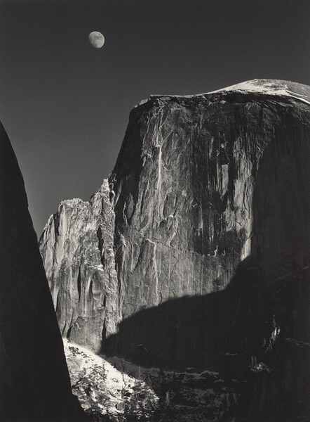 From a low vantage point, we look up at the rugged face of a tall, almost sheer rock formation with the moon above in this vertical black and white photograph. Filling most of the composition, the rock formation is slightly rounded along the top but dips down to a jagged ridge to our left. Strong light from the upper right creates deep shadows across its iron-gray surface, which is textured with long vertical streaks of white and lighter gray. An almost-full moon hovers in the otherwise clear, charcoal-gray sky above it, near the top of the image to our left of center. Formations outside our view cast deep shadows along the right edge of the photograph. A smattering of white across from us, low in the image, suggest snow on the rock there. A dark mass close to us is an ink-black silhouette rising from the lower left corner and angling off the left edge, near the upper left corner.