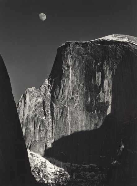 From a low vantage point, we look up at the rugged face of a tall, almost sheer rock formation with the moon above in this vertical black and white photograph. Filling most of the composition, the rock formation is slightly rounded along the top but dips down to a jagged ridge to our left. Strong light from the upper right creates deep shadows across its iron-gray surface, which is textured with long vertical streaks of white and lighter gray. An almost-full moon hovers in the otherwise clear, charcoal-gray sky above it, near the top of the image to our left of center. Formations outside our view cast deep shadows along the right edge of the photograph. A smattering of white across from us, low in the image, suggest snow on the rock there. A dark mass close to us is an ink-black silhouette rising from the lower left corner and angling off the left edge, near the upper left corner.