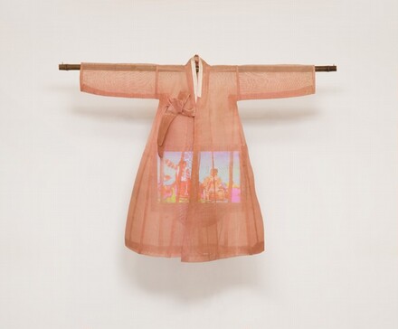 A lightweight, sheer, coral-peach robe hangs from a wooden dowel over a color television screen in this sculptural piece, which hangs against a white wall. The robe has long sleeves extending horizontally along the dowel, which extends a bit past the cuffs. The robe has a narrow, cream-white collar and ties to one side over the chest. It flares slightly below the arms, creating a bell shape. The rectangular video screen that hangs within is partially obscured, but looks like at least two people in a brightly colored setting.
