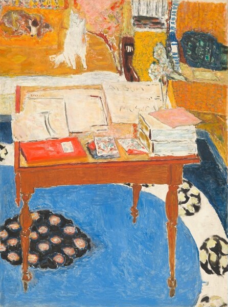We look down onto papers and books piled on a small wooden table with delicate, spindle legs, with a dog and cat on a sofa in the background in this vibrantly colored, stylized, vertical painting. The scene is loosely painted with dabs and visible brushstrokes in butterscotch and golden yellow, azure and navy blue, apple red, and white. The square table takes up most of the bottom two-thirds of the composition. Its surface is almost entirely covered with a spread of papers and stacks of books, including two thick, white volumes near the front, right corner and a thin red book on the other front corner. Smaller squares between the books are painted with streaks and washes of turquoise blue, scarlet, and brick red. A tall, narrow vase with abstracted, ice-blue flowers sits behind the stacked books, to our right. Beyond that, at the back right corner of the table, a sheet of paper reads “ALBUM MUSICAL,” with the final L partially obscured by a piece of bubblegum-pink paper set askew atop the stacked books. The artist’s name appears in rust-red letters on a paper at the back left corner of the table: “Bonnard.” The rug beneath the table is dominated by a vivid, azure-blue ring. The blue ring is surrounded by a white band decorated with widely spaced, black ovals dotted with pale lemon yellow. At the center of the blue ring is a black circle dotted with abstracted rosettes made with rings of apricot orange and gray around yellow centers. At the back edge of the rug, at the top left corner of the painting, a white cat sits and brown spotted dog lies on a mustard-yellow sofa, which has a wooden arm on the end we can see. There is a tangerine-orange chair next to it, in the upper right corner of the painting, and a teal-blue block there could be a pillow. The floor between the furniture is goldenrod yellow.