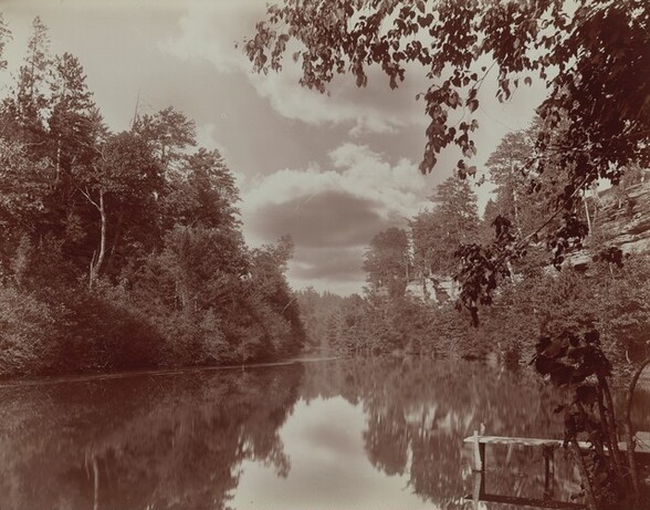 We look across a glassy lake at a V-shaped slice of cloudy sky between banks of trees that reach from the top corners of this horizontal black and white photograph to just below the center of the composition. The forest and sky are reflected in the water below, turning the V of sky into an X. A wooden dock extends into view from the lower right corner, and a few branches from a tree out of sight dip down across the right half of the top edge.