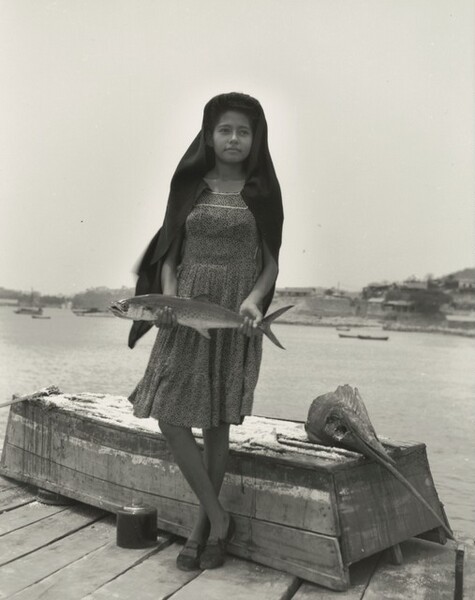 A dark-skinned girl stands on a dock holding a fish wider than her hips down in front of her with both hands, one ankle crossed over the other in this black and white photograph. Her body faces us, but she looks up and off to our right. A dark cloth covers her hair and shoulders over a knee-length dress. She wears loafer-style shoes. She stands in front of an upturned rowboat with a severed swordfish head laid on the flat stern, to our right. The long sword-like upper lip angles into the lower right corner of the photograph, and the fish’s large eye is a gaping hole. Buildings on a spit of land that comes about halfway up the composition enclose the body of water in the distance. The contrast of the girl’s dark head covering against the white sky creates an optical effect, making the cloth seem to glow.