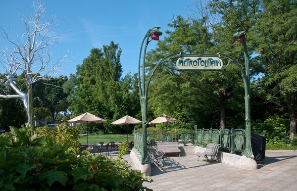 Set in an outdoor garden, a sculpture with two tall metal arms curve up like stylized tree branches to hold a sign between them, which reads, “METROPOLITAIN.” In this photograph, the sign faces us and fence-like rows of panels extend back from the posts, and across the back to enclose three sides. The panels, posts, and the framework holding the sign are all painted sage green. Each panel has a rounded top with a two-lobed, medallion-like center. The arms of the posts reach above the sign, and each curves up and over a burgundy-red light that hangs down like a flower bud. Three wooden benches sit within the enclosure. The sculpture is set within a garden with tall trees, a grassy lawn, and umbrella-covered café tables and chairs. A silver, metal tree rises in the near distance to our left.