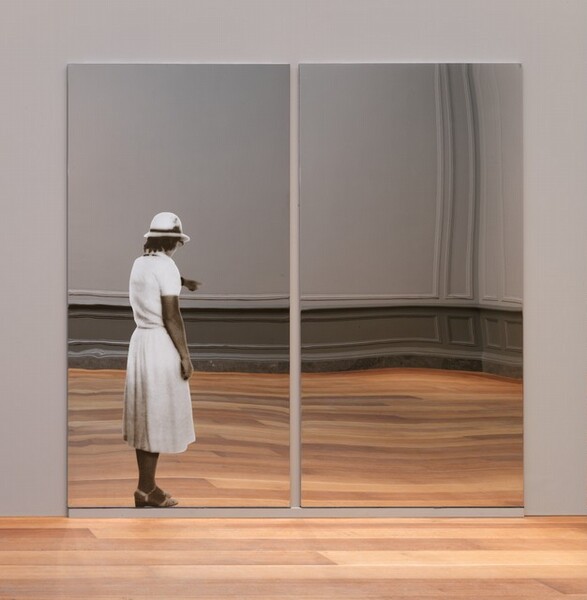 Two vertical, rectangular, polished steel panels are hung close to each other on a dove-gray wall, and the wavy reflections show the wood floor and gray wall behind us. In the left panel, the image of a woman is silkscreened onto what appears to be a mirror. The woman has brown skin, and she stands facing away mostly away from us. She points into the distance with her left hand, farther from us, and her other arm hangs by her side. She wears a rounded white cap with a narrow brim over shoulder-length dark hair. The edge of her glasses are visible over her right cheek. Her white dress is cinched at the waist, has short sleeves, and comes to mid-calf. She wears white sandals with low heels. The room reflected around her has dark gray paneling along the bottom of the lighter gray walls.