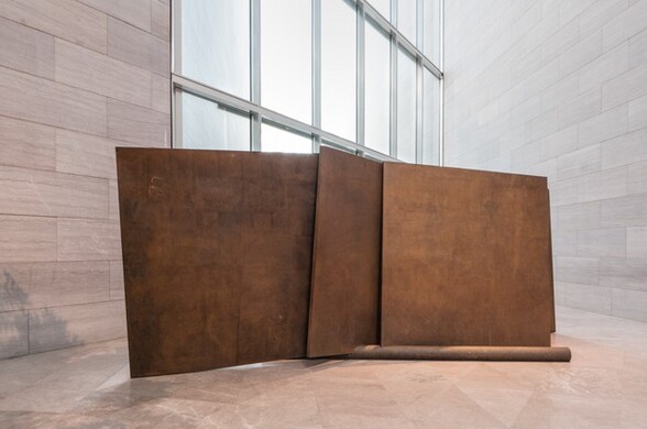 This free-standing, abstract sculpture is made of five square or rectangular bronze-colored plates braced by two long poles that lie on a pale pink marble floor. In this photograph, we see one pole at the foot of three of the plates, which lean against each other at different angles. The sheets and poles are mottled with bronze and brown, and the surface is textured, as if it would be a little rough to the touch. At least two of the plates sit in grooves notched into the pole on the floor. In our view, the sculpture sits in front of a two-story bank of windows, which continue off the top edge of the photograph. The sculpture almost entirely blocks the lower story of windows. The walls to either side of the windows meet at an acute angle along the right edge of the windows. The walls and floor are both covered with pale pink marble.