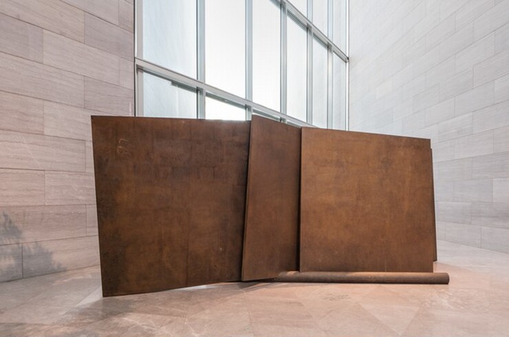 This free-standing, abstract sculpture is made of five square or rectangular bronze-colored plates braced by two long poles that lie on a pink marble floor. In this photograph, we see one pole at the foot of three of the plates, which lean against each other at different angles. The sheets and poles are mottled with bronze and brown, and the surface is textured, as if it would be a little rough to the touch. At least two of the plates sit in grooves notched into the pole on the floor. In this view, the sculpture sits in front of a two-story bank of windows, which continue off the top edge of the photograph. The sculpture almost entirely blocks the lower story of windows. The walls to either side of the windows meet at an acute angle along the right edge of the windows. The walls and floor are a pale pink marble.