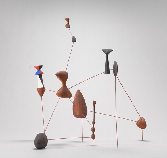 This sculpture is made up of ten shapes connected with rigid, straight brown wire to create an interlocking, web-like formation that rests on the floor. Five of the shapes appear to be raw wood and the other five are painted. Four of the painted shapes are dark gray. The fifth painted piece, at the upper left in this photograph, has a wooden cone-shaped tip pointing downward beneath a black and white mid-section, and an angular, hourglass-shaped tail painted in triangular blocks of red, blue, and black. The other wooden and painted shapes range in size and form, with an almond-shaped piece at the center, rounded pieces throughout, and two rounded hour-glass shaped pieces near the upper left.