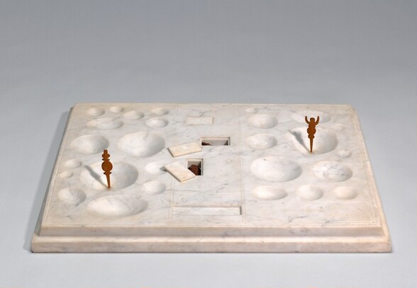 Carved from a slab of white marble, the top surface of this sculpture is covered with shallow, cuplike and deep rectangular depressions of various sizes. We look slightly down onto the slab in this photograph. The surface is divided into three vertical sections, which are defined by incised lines. The cuplike depressions are in the sections to the left and right. Two tiny, carved, dark wooden objects, like game pieces, stand upright in a cup with one the left and one to the right. Each piece is flat with a symmetrical design and a point at the bottom that inserts into the cup. The left piece has a disk flanked by triangular protrusions with a clover shape at top. The second piece has four stacked, small circles, and a U-shaped form to suggest the outline of a person with raised arms. Three smaller but deep rectangular cavities are carved into the middle section, and each one has a lid. Two lids sit askew on the top surface of the board near their respective openings, and inside are additional wooden pieces. A blank, rectangular section outlined with lightly inscised lines in the lower right is carved with reversed script letters that read, “on ne joue plus.” The entire surface has beveled edges, so it sits on a stepped base.