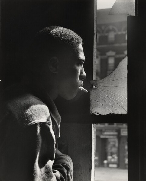 A young Black man stands in profile looking out a tall window to our right in this vertical black and white photograph. The image is cropped so his head, shoulder, and upper arm fill the left half of the composition. He has short hair and wears a shirt or jacket that diffuses the light to suggest that it could be flannel or another soft fabric. With a cigarette dangling loosely from his lips, he stares through the shattered upper pane of the window and he holds his right hand across his chest. The strong light source from the right accentuates his nose, cheeks, hair, and shoulder, and the space behind him is lost in shadow. A blurred, three-story building across the street has a dark façade with white stone lintels above the windows and a mansard roof with curved dormers.