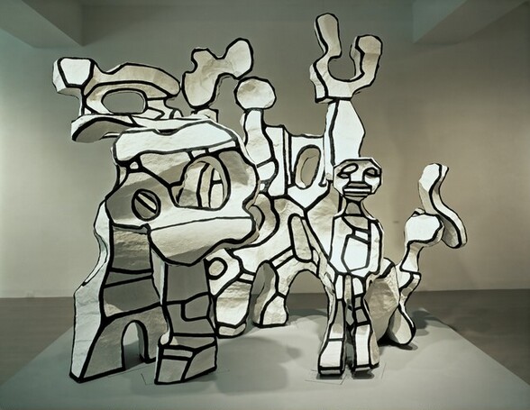 Abstract forms in white, outlined with bold black lines, turn, twist, and curve up around a person with cartoon-like features in this free-standing sculptural piece. The shapes create legs and are pierced with holes, like an abstracted coral reef. The forms are bright white with thick black outlines around all the edges and crossing the curves and angled sides of the forms. In this photograph, it is not possible to see if a vaguely H-shaped section to our left is attached to or stands independently from the row behind it. A person with abstracted eyes, nose, and mouth stands in front of the sculpture, to our right. In this photograph, the sculpture stands on a plinth in a room with a wood floor and a white wall behind it.