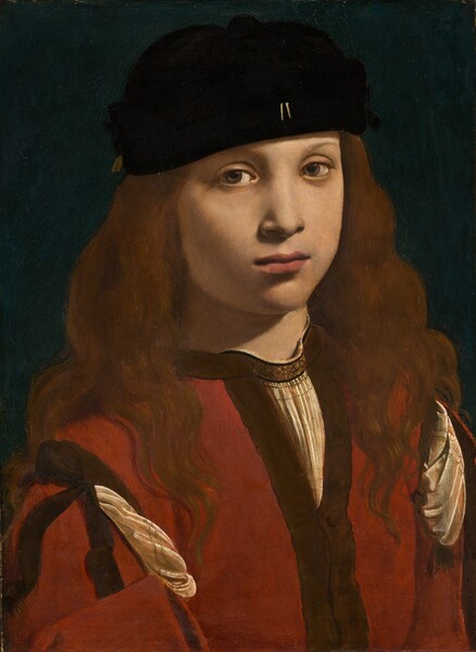 The head, shoulders, and chest of a light-skinned boy or young man wearing a black cap and red garment nearly fill this vertical portrait painting. His body is angled slightly to our right, and he looks at us from the corners of his hazel eyes. He has smooth skin, rounded cheeks, faint, thin eyebrows, and a small nose. His full, dark pink lips are closed. He wears a brimless, soft black cap, slightly askew, over long chestnut-brown hair that covers his ears and flows down past his shoulders. Two small, gold, vertical lines accent the front center of his cap near the lower edge. He wears a collarless, crimson-red jacket, with dark brown trim along the edges and at the shoulder. Pleated folds of his ivory-white shirt material puff out from openings at the shoulders and a V at the top of the chest. There is a also brown band around the neck of the white shirt with a pattern of black, knot-like designs. The white shirt also has two rows of delicate black lines where it is pulled through at the shoulders. The boy is lit from our right and above, casting a shadow on his neck and cheek to our left. The background behind him is teal blue.