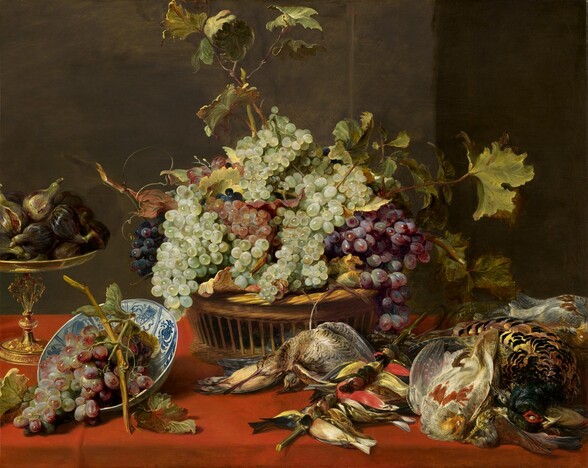 A basket overflowing with grapes and surrounded by dead birds and more fruit sits on a long table covered in a scarlet-red cloth in this horizontal still life painting. In the basket at the center of the painting, luminous green, blue-black, red, and purple grapes are piled up in a rough pyramid that overflows the sides. Long stems with broad sage and celery-green leaves edged with brown jut up above and to the side of the bunches of grapes, nearly touching the top edge of the painting. Just in front of the basket to our left, a bowl overflowing with a bunch of purple grapes is tipped toward us. The inside of the bowl is painted with cobalt-blue, geometric, vegetal designs against a white background. Behind it, at the far left, a tall, bronze, footed serving dish stacked high with pale green and black-skinned figs is cut off by the left edge of the composition. In front of and to the right of the central basket, the bodies of about two dozen small and medium-sized birds fill the right half of the tabletop. About ten small birds are arranged along a stick between a pair of dead birds to our left and a pile of several larger birds to our right. The birds’ feathers and bodies are painted in shades of cream and parchment white, ink black, ruby red, tawny brown, and butter and golden yellow. The fruit and birds are lit from the upper left, and the backround is earth brown.