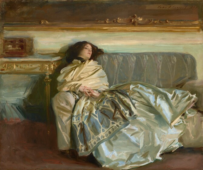 A woman with pale skin and dark brown hair is swathed in silky fabric as she reclines along a gray couch in this horizontal painting. Her head rests along the backrest of the sofa to our left, so her body extends to our right. Her torso is wrapped in a voluminous, ivory-white scarf or wrap. The scarf wraps tightly around her neck, and the bottom edge, near her knees, has a wide, indigo-blue pattern of ovals and vegetal forms. Her shimmering silver gray skirt is painted loosely with baby and sky blue, olive and sage green, and white strokes, and it drapes over the cushions and down the front of the couch. Her hands are clasped so her interwoven fingers rest at her navel. She looks down and off to our right. A table to our left is edged with gold. The wall behind her, above the couch, is painted with long streaks of eggshell white and pale turquoise. The gold frame of a dark painting hanging over the couch spans the width of the composition. Near the upper right corner of the canvas, the artist included a signature and as if he had signed the painting within this painting with loose, dark letters: “John S. Sargent 1911.”