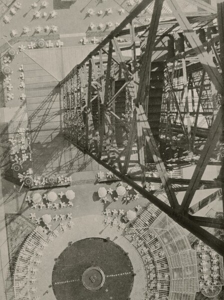 From very high up, we look down along the height of a metal radio tower at a plaza on the ground, which is dotted with umbrellas, tables, and chairs in this vertical black and white photograph. The side of the tower we look down from fills almost the height of the right edge of this image. Crisscrossing girders form diamond shapes down the sides, enclosing stairways and other structures. In the plaza below, the tables and chairs create cross and asterisk shapes around the discs of the umbrellas, seen from above. Along the bottom of the photograph, and cut off by that edge, the tables and a skeletal awning creates a ring around a circular area, presumably grass.
