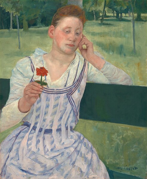 Shown from the knees up, a young woman with peachy skin and red hair sits facing us on a pine-green bench in this vertical painting. She sits with her body angled to our right, and her left elbow, on our right, hooks over the top slat of the bench. Her head tilts slightly to our right to rest on the curled fingers of that hand. In her other hand, she holds up a red flower on a short stem. Her hair is pulled up and back over a high forehead, and her cheeks are deeply flushed. Her pink lips are closed, and she looks down and to our left at the flower with gray eyes. She has an oval-shaped face and a hint of a double chin. Her white dress is painted with ice-blue shadows and folds. It has a deep V neck that reaches to an apron-like pinafore patterned with vertical bands of pale lilac that alternate with angled daubs of blue. Beyond her is a loosely painted landscape with grassy areas and clusters of tall trees painted in spring, emerald, and sea glass green. The edge of the grassy field nearly brushes the top edge of the canvas, and the trees are cropped by it. The artist signed the lower right, “Mary Cassatt.”
