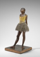 Sculpted with rich brown wax, a young ballerina stands with her arms straight, hands clasped behind her back, and one foot in front of the other on a square wooden base. Her body is angled to our left in this photograph. Both feet are splayed outwards and her right foot is placed far in front of her left. Bangs cover her forehead, and she has a heart-shaped, upturned face with a squat nose and slightly pursed lips. Her heavy-lidded eyes are nearly closed and her hair is pulled back and tied with a wide, cream-colored ribbon. She wears a fabric costume with a sleeveless, gold-colored bodice, a gray tulle skirt, and ballet slippers. Her body is sculpted from dark brown wax and a layer of wax covers her hair, bodice, and ballet slippers. 