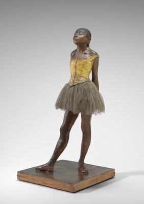 Sculpted with rich brown wax, a young ballerina stands with her arms straight, hands clasped behind her back, and one foot in front of the other on a square wooden base. Her body is angled to our left in this photograph. Both feet are splayed outward, and her right foot is placed far in front of her left. Bangs cover her forehead, and she has a heart-shaped, upturned face with a squat nose and slightly pursed lips. Her heavy-lidded eyes are nearly closed and her hair is pulled back and tied with a wide, cream-white ribbon. She wears a fabric costume with a sleeveless, gold-colored bodice, a gray tulle skirt, and ballet slippers. Her body is sculpted from dark brown wax, and a layer of wax covers her hair, bodice, and ballet slippers.