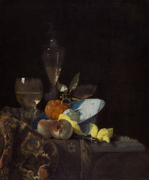 A blue and white porcelain bowl, fruit, wine glasses, and a woven rug are gathered on a tabletop in front of a black background in this vertical still life painting. Light strongly illuminates the objects, creating a stark contrast to the dark background. At the center of the composition, the porcelain bowl is tipped so its wide brim rests on the rug bunched on the tabletop to our left. The rug is painted with a pattern in earthy beige and chocolate brown, denim blue, bronze, and cream white. A piece of roughly textured orange fruit resembling a tiny pumpkin has a stem with a small white blossom growing from its top. The fruit sits in the bowl alongside a vivid yellow lemon that has been partially peeled. The top has been cut from the lemon to form a long strip that curls onto the table, and the pulp glistens in the light. Two peaches sitting in front of the bowl are painted with tones of silvery ivory and burnt orange. The handle of a utensil juts out from between the peaches and lemon peel. Two glasses and a slender carafe are arranged in a row behind the fruit and bowl. The glass to the left has a round bowl over a thick, columnar stem, and the tall, gently flaring glass carafe behind the orange fruit are both half-filled with pale gold liquid. A drinking glass with a wide, shallow, scalloped-edged cup is mostly hidden behind the porcelain bowl, to our right.