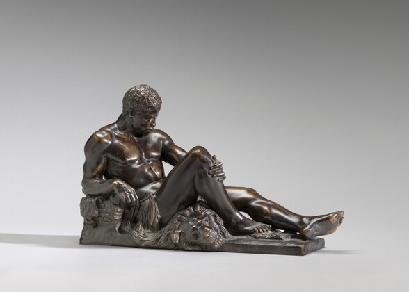 A muscular, nearly nude Black man reclines on a lion’s skin in this freestanding bronze sculpture. In this photograph, his head is to our left and his feet extend to our right. His face tips down, and he has short, curly hair, a furrowed brow, and a small, rounded nose. His full lips are closed, and he has hollows in his cheeks. He leans onto his right elbow, closer to us, which rests on an object, perhaps a basket. His right knee is raised so that foot is flat on the ground. He clutches the bent leg just below the knee with his other hand, and his left leg stretches long. The lion’s head, with eyes closed, rests beneath the bent knee. Long fur, hair, or another material hangs from a rope around the man’s waist. He and the pelt rest on a narrow rectangular base. The dark patina of the bronze has been burnished or rubbed gold in some areas, especially along the ridges of the man’s muscles and on the extended foot.