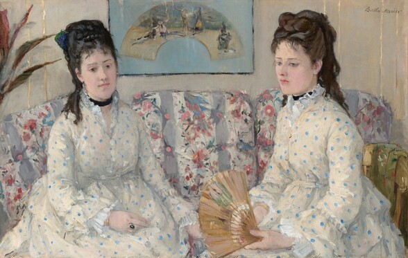 Shown from the lap up, two women with pale skin and dark hair pulled up and back, wearing white dresses with baby-blue polka dots, sit on a couch in this horizontal portrait painting. Both women have straight, dark brows, delicate noses, smooth skin, and their small, rose-pink mouths are closed. Wispy bangs brush their foreheads, and their hair is piled high with long ringlets falling down their backs. Both wear black ribbons like chokers around their necks. Their dresses have high necks with ruffles along the necklines, ruffles at the wrists, and are gathered under the bust. They sit angled in toward each other. The woman on our left has black hair, and she looks down and to our right with dark eyes. She wears a gold ring with a dark, oval stone on one hand resting in her lap. The other woman has chestnut-brown hair and looks down and off to our left with ice-blue eyes. She holds an open fan in her hands in her lap. The fabric on the couch has vertical white and lilac-purple stripes, and is overlaid with a pattern of pink flowers and green leaves. A framed picture hanging on the bone-white wall behind the women shows an arched painting against a sky-blue background. A few spiky leaves from a houseplant are cut off by the left edge of the painting. The portrait is loosely painted throughout, especially in the couch and background. The artist signed the painting in the upper right corner, “Berthe Morisot.”