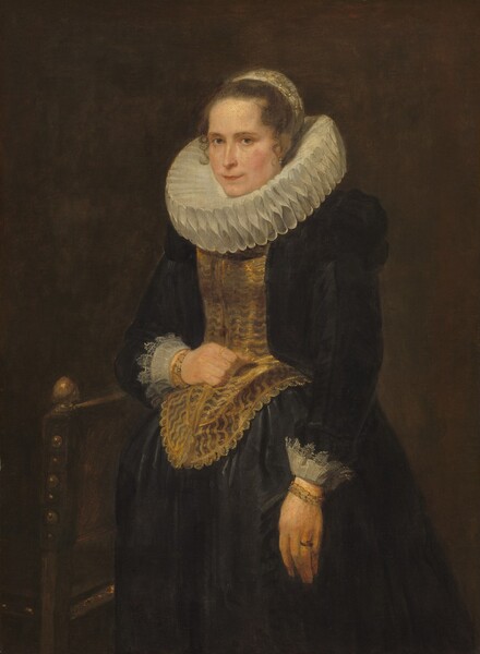 Shown from the knees up, a pale-skinned woman wears a wide, pleated collar and a black gown in this vertical portrait painting. Her body is angled to our left, and she looks at us from the corners of her light brown eyes. She has low brows, a straight nose, and her thin lips are closed. Her hair is pulled back under a pearl-white headdress at the back of her head, and a few tendrils curl alongside her face. Pearls hang from the ear we can see. The figure-eight pleats of the collar are as tall as her neck so her chin rests on it, and it is nearly as wide as her shoulders. A gold-colored corset comes down in a long U shape over her belly. Her black dress has puffed shoulders, and white lace extends back from the cuffs of the long sleeves. The full, black skirt flares out from her waist. She wears gold bracelets on both wrists. Her left arm, to our right, hangs down in front of her, and she wears a ring on the third finger of that hand. The back of a chair is at her other elbow, which is bent so she can rest a loose fist against her corset. She wears a gold ring on that index finger. The sides of the back of the chair are studded with brass bosses, and a gold-colored finial is at the front. The background is earthy brown.