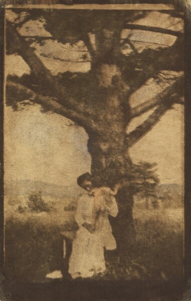 A mother sitting in front of a tree braces a naked child who stands on her lap in this vertical photograph. The print is made with multiple exposures, some in faint peach or slate blue, which creates a blurred, ethereal effect. The tree’s thick trunk rises up and off the height of the photograph. The woman wears a white dress, and her dark hair is tied up. She leans her head down, perhaps tucking her chin into the child’s soft elbow. The child faces away from us, the dark hair blending into the tree trunk. A meadow beyond the tree leads back to hazy mountains, which come about a third of the way up the composition, under a clear sky.