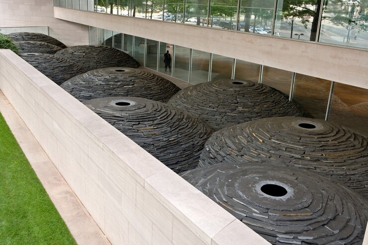 We look down onto nine gray stone domes that melt into each other in the space of a patio enclosed with a pink marble wall opposite floor-to-ceiling windows. Each dome is nearly as tall as the ceiling over the windows, and each is made of stacked pieces of slate. At the top center of each dome is a collar of gray slate to create a pupil-like opening. The stone collar creating the openings is lighter gray around the ink-black centers, which are the shaded interiors of the domes. The domes meld and stack like a handful of bubbles.