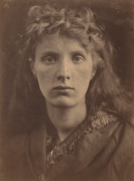 The head and shoulders of a young woman with pale skin and long hair nearly fill this vertical, sepia-toned photograph. Shown against a dark background, she looks directly at us. Her nose is straight and her lips closed. Wavy curls piled over her forehead suggest that her hair is partially pulled back or pinned up around her oval face, and it falls behind her shoulders. Her garment has wide lapels and is worn over patterned clothing.