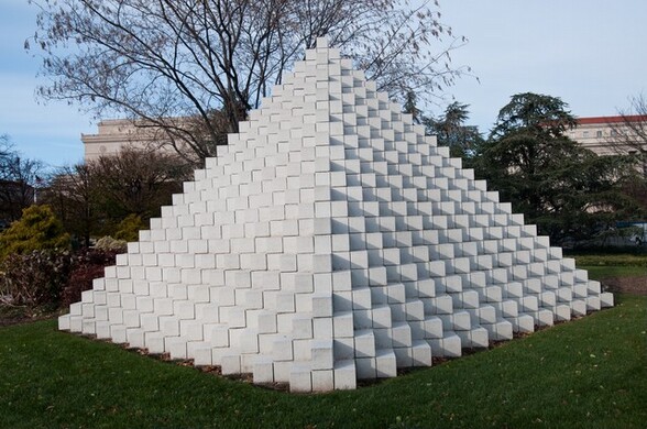 A stepped pyramid made of interlocking, white concrete blocks sits on a grassy lawn, rising above the bushes and low trees behind it in this photograph. This view shows the pyramid with one corner coming straight at us so each side is a zigzag of twenty-four steps. The structure is lit from our left so casts gray shadows to our right. One tall tree behind the pyramid extends off the top edge of the photograph and buildings in the distance have cream-white walls and red roofs.
