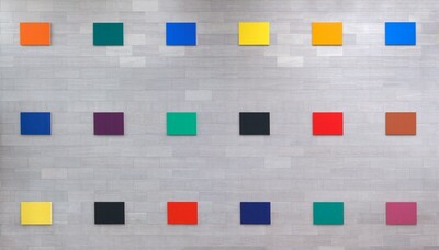 Three rows of six horizontal, rectangular canvases, each painted a single, saturated color, hang in a widely spaced grid against a three-story gray stone wall in this photograph. Moving from our left to right, the canvases in the top row are carrot orange, pine green, royal blue, canary yellow, marigold orange, and azure blue. The middle row has panels in marine blue, amethyst purple, shamrock green, soot black, vivid red, and rust brown. The bottom row canvases are banana yellow, black, coral red, midnight blue, emerald green, and orchid purple. The smaller, rectangular stones in the wall behind the panels are also laid horizontally, and they vary from silvery white to smoke gray.