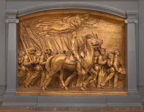 A white man in military uniform rides a horse in front of a regiment of five rows of Black troops in this sculpture, which is painted entirely in gold. The artist created a shallow, stage-like space with an arched top so the men are sculpted in three dimensions, though they become more compressed as they move back in space. The men and horse face our right in profile in this view. The man on the horse has a pointed, straight nose and a goatee. He wears a cap with a flat top and narrow brim, a knee-length coat, gloves, and knee-high boots with spurs. He holds a thin sword down by the side of the horse with his right hand and holds the reins of the horse with his left. The horse’s head is pulled upwards by the short reins and its mouth is open around the bit. About twenty soldiers are lined up in rows beyond the horse, and they march in unison. They carry blankets rolled atop knapsacks, canteens, and rifles resting on their right shoulders. However, the details of how their uniforms bunch up around their equipment and the way their caps have been molded and fit is unique to each person. Their ages also vary from young, cleanshaven individuals to bearded older men. Two men carry furled flags near the back, to our left, and a drummer boy plays at the head of the regiment, to our right. All the men look straight ahead, their lips closed. A female figure in a billowing robe floats above them under the arched top with her eyes closed. Her left arm is outstretched and she holds a laurel branch and poppies close to her body with her right arm. An inscription in the upper right corner is created with raised capital letters: “OMNIA RELINQVIT SERVARE REMPVBLICAM.” A longer inscription is carved into the base along the bottom edge of the memorial, also in all caps: “ROBERT GOVLD SHAW KILLED WHILE LEADING THE ASSVLT ON FORT WAGNER JVLY TWENTY THIRD EIGHTEEN HVNDRED AND SIXTY THREE.” The artist’s signature is inscribed In the lower right corner, in smaller letters: “AVGVTVS SAINT GAVDEN M-D-C-C-C-L X X X X V I I I.”