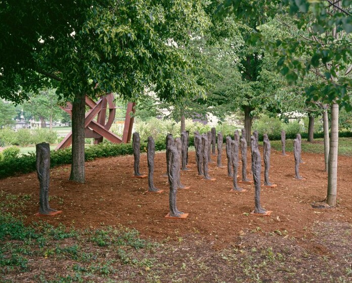 Twenty-one, free-standing, bronze sculptures of headless people are arranged under the green canopies of trees in this horizontal photograph. The earth-brown surfaces of the sculptures are rough. In this view, they cluster together at various distances from each other, and the bodies of all are angled to our right. They stand erect with their legs together and arms pressed tightly along their sides. From this angle, one person is positioned farther from the rest, to our left. Each person stands on a square base that sits on the ground, which appears to be fine mulch or pine needles. Six trees are spaced around the group, and together their canopies fill the top third of this photograph. Beyond the trees is a low, green hedge that encloses the whole group. Sunlight illuminates the front, right-hand surfaces of the sculptures.