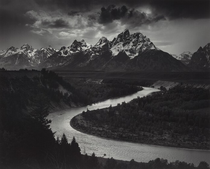 We look down into a darkened valley with a shimmering, curving river winding back to a screen of craggy, snow-topped mountains in the distance in this horizontal black and white photograph.