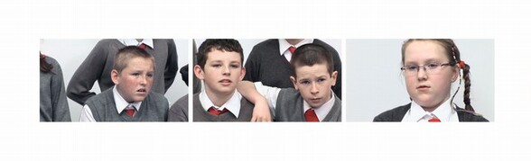 Three still images from a video installation show the faces of four pale-skinned age children wearing school uniforms against white backgrounds. The uniforms consist of white dress shirts, red ties, and gray vests or sweaters. From left to right, the first image shows a boy gaping, mouth open. The middle picture shows two boys, one of whom slings an arm onto the other’s shoulder. One looks steadily on, the other with brows furrowed. The image to our right shows a girl wearing glasses, her hair in thin braids. She also looks steadily at something out of our view. The shoulders and elbows of students sitting next to or standing behind those we see are visible. The three images are printed close together in a row, like a sideways piece of film.