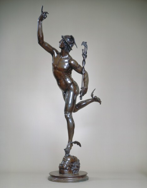 A nude, muscular man with wings on his ankles balances on the toes of one foot as the other leg and one arm raise up, creating a spiral that moves from the toes of his weight-bearing foot to the top of his opposite, raised hand in this freestanding bronze sculpture. In this photograph, his body is angled to our left and he looks off over his right shoulder, farther from us, so we see his face in profile. He wears a smooth, dish-like helmet with wings to each side over short, curly hair. He has a long, straight nose, and his lips are parted. His right arm, farther from us, reaches high into the air with his index finger pointing upward. He holds a scepter tucked near his side with his other hand, closer to us. The staff is as tall as the man’s torso, and has two snakes entwined up its length with wings at the top. His right leg is raised behind him as he leans a bit forward over the other toes, which balance on a narrow wedge. The wedge is held in the mouth of a human head tipped back so it rests on the disk-like base.