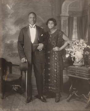 An elegantly dressed Black man and woman stand facing and looking at us, slightly smiling, in a room in this vertical photograph. The image is monochromatic like a black and white photograph but is printed in warm tones of golden and dark browns. To our left, the man has short-cropped hair and is cleanshaven. He wears a three-piece tuxedo and holds a bowler hat and cane in his right hand, on our left. He pulls his suit jacket back to hook his opposite thumb in his vest pocket. He wears rings on each of his pinky fingers and a chain crosses his vest, tucked into the same pocket as his thumb. The woman stands with her left hand, on our right, on her hip and her opposite hand resting on the man's shoulder. She has a cheek-length bob haircut and wears dangling earrings and a necklace with a pendant. Her ankle-length, sleeveless dress is beaded with geometric and scrolling patterns. Some of the beads and the ring she wears on the fourth finger of the hand on her hip catch and reflect the light. An upholstered chair sits to our left and a wood side table with an urn filled with flowers and a telephone stands to our right. The telephone has a conical earpiece hanging from a stand with the flaring mouthpiece. The background behind the people has a painted or wallpapered section to our left and an arch leading to a curtained window to our right. Parts of the photograph are noticeably out of focus, particularly the background and flowers. The artist signed the work with white letters against the dark shadows under the seat of the chair, near the lower left corner: 