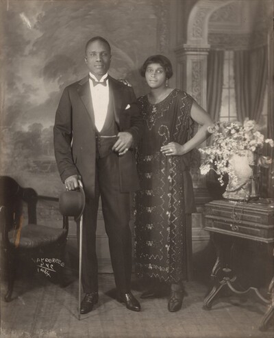 An elegantly dressed Black man and woman stand facing and looking at us, slightly smiling, in a room in this vertical photograph. The image is monochromatic like a black and white photograph but is printed in warm tones of golden and dark browns. To our left, the man has short-cropped hair and is cleanshaven. He has dark eyes, a rounded nose, and his lips are closed in a slight smile. He wears a three-piece tuxedo and holds a bowler hat and cane in his right hand, on our left. He pulls his suit jacket back to hook his opposite thumb in his vest pocket. He wears rings on each of his pinky fingers and a chain crosses his vest, tucked into the same pocket as his thumb. The woman stands with her left hand, on our right, on her hip and her opposite hand resting on the man’s shoulder. She has a delicate nose, and also has dark eyes and her closed lips turn up slightly at the corners. She has a cheek-length bob haircut and wears dangling earrings and a necklace with a pendant. Her ankle-length, sleeveless dress is beaded with geometric and scrolling patterns. Some of the beads and the ring she wears on the fourth finger of the hand on her hip catch and reflect the light. An upholstered chair sits to our left and a wood side table with an urn filled with flowers and a telephone stands to our right. The telephone has a conical earpiece hanging from a stand with the flaring mouthpiece. The backdrop behind the people has a painted or wallpapered section to our left and an arch leading to a curtained window to our right. Parts of the photograph are noticeably out of focus, particularly the background and flowers. The artist signed the work with white letters against the dark shadows under the seat of the chair, near the lower left corner: “VAN DER ZEE NYC 1924.”
