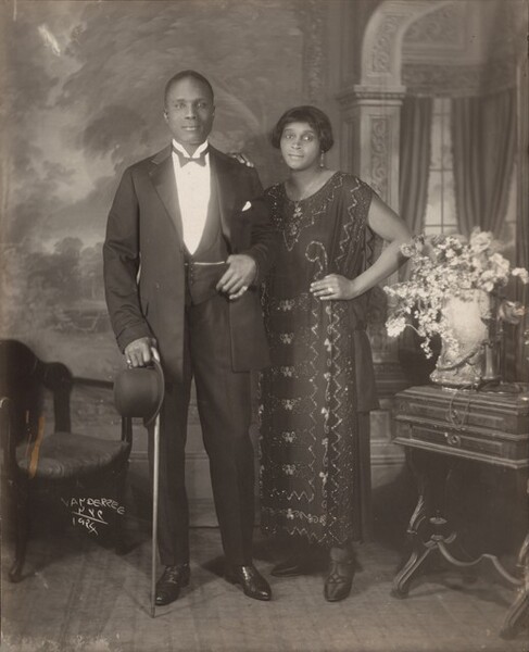 An elegantly dressed Black man and woman stand facing and looking at us, slightly smiling, in a room in this vertical photograph. The image is monochromatic like a black and white photograph but is printed in warm tones of golden and dark browns. To our left, the man has short-cropped hair and is cleanshaven. He has dark eyes, a rounded nose, and his lips are closed in a slight smile. He wears a three-piece tuxedo and holds a bowler hat and cane in his right hand, on our left. He pulls his suit jacket back to hook his other thumb in his vest pocket. He wears rings on each of his pinky fingers and a chain crosses his vest, tucked into the same pocket as his thumb. The woman stands with her left hand, on our right, on her hip and her other hand resting on the man’s shoulder. She has a delicate nose, dark eyes, and her closed lips turn up slightly at the corners. She has a cheek-length bob haircut and wears dangling earrings and a necklace with a pendant. Her ankle-length, sleeveless dress is beaded with geometric and scrolling patterns. Some of the beads and the ring she wears on the third finger of the hand on her hip catch and reflect the light. An upholstered chair sits to our left and a wood side table with an urn filled with flowers and a telephone stands to our right. The telephone has a conical earpiece hanging from a stand with a flaring mouthpiece. The backdrop behind the people has a painted or wallpapered section to our left and an arch leading to a curtained window to our right. Parts of the photograph are noticeably out of focus, particularly in the background and the flowers. The artist signed the work with white letters against the dark shadows under the seat of the chair, near the lower left corner: “VAN DER ZEE NYC 1924.”