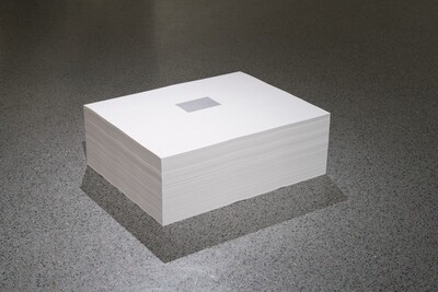 We look down onto a tidy stack of hundreds of sheets of white paper, which sits on a gray, speckled floor in this photograph. The top sheet is printed with a small, dove-gray rectangle at the center. A corner of the stack angles toward us, with the short side of the sheets to our left and the long side to our right.